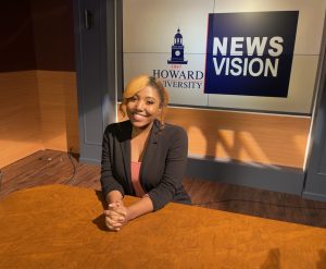 Ianna Fenton, a young woman with blonde hair, wearing a black blazer, smiles while seated at a news anchor desk with a "Howard University Newsvision" logo in the background.