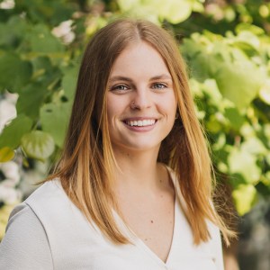 Headshot of Abbey Gingras. She is pictured with long, straight blonde hair and a bright smile, wearing a white top, standing in front of lush green foliage.