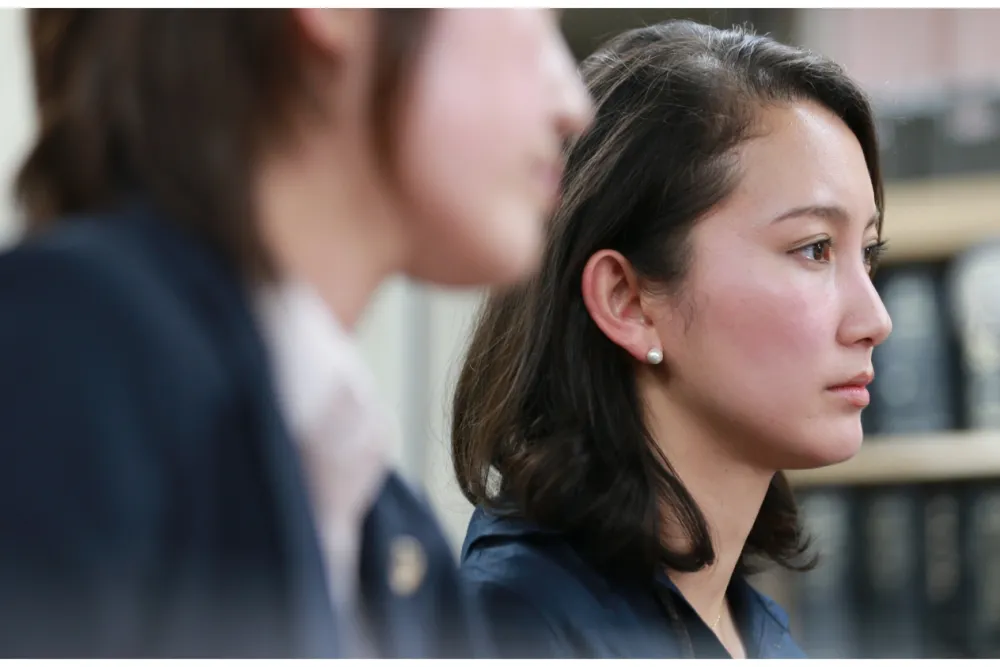 Two women in a professional setting are seated and focused. Shirori Itō, the filmmaker and subject of BLACK BOX DIARIES, is the woman in the foreground. She has shoulder-length dark hair, wearing pearl earrings and a blue blazer. The background is slightly out of focus, suggesting an office or court environment.