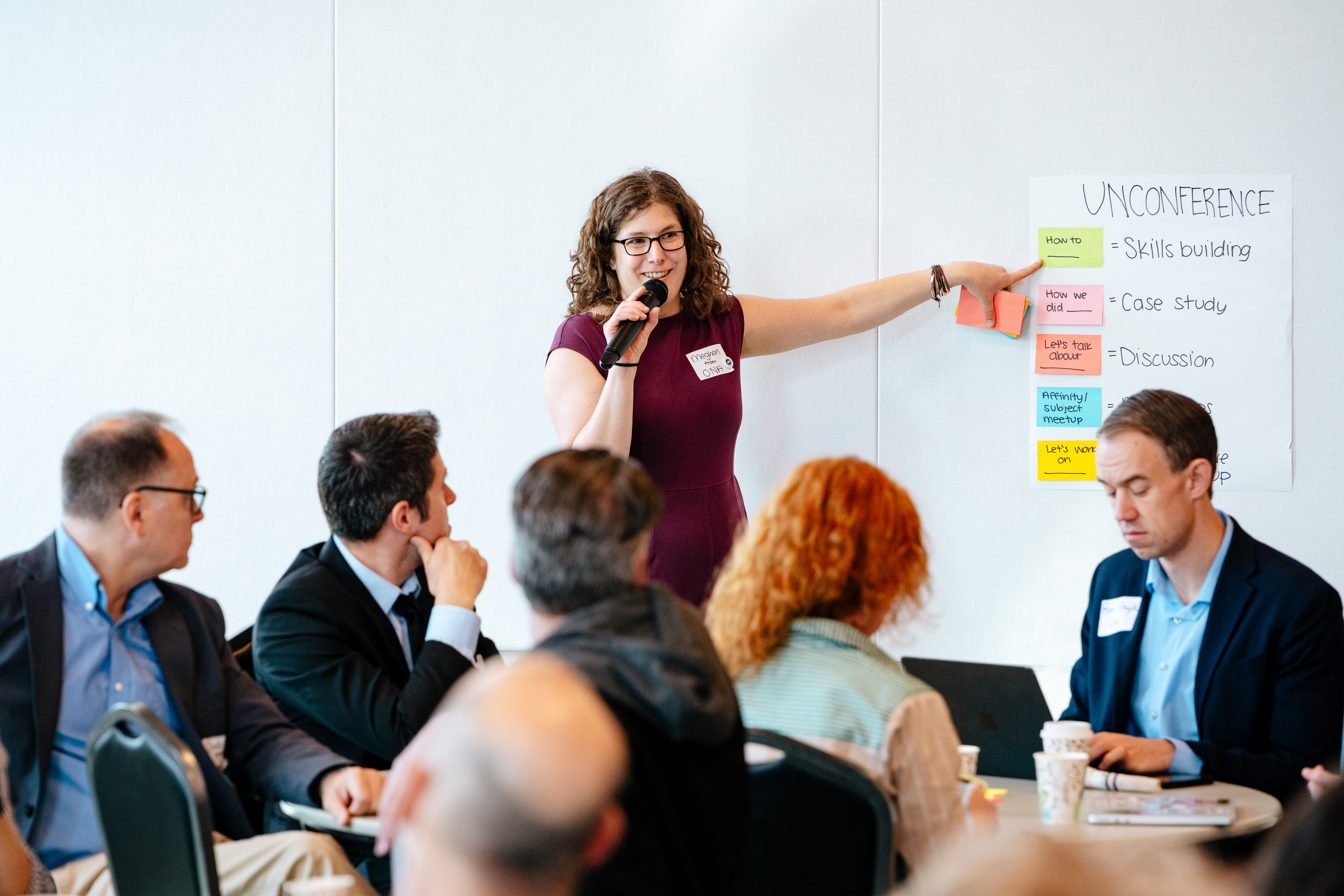 Meghan Murphy stands in front of a crowd of people holding a microphone and pointing to a sign labeled UNCONFERENCE, with different colored post-it notes on it.