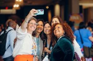 Five women standing closely together look up, smiling at a cell phone held by the left-most woman to take a group selfie