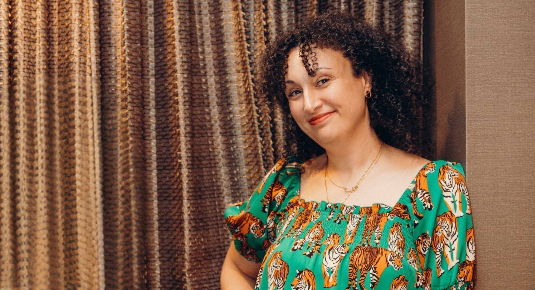 Hanaa stands smiling leaning against a wall. She is wearing a green dress with a tiger print.